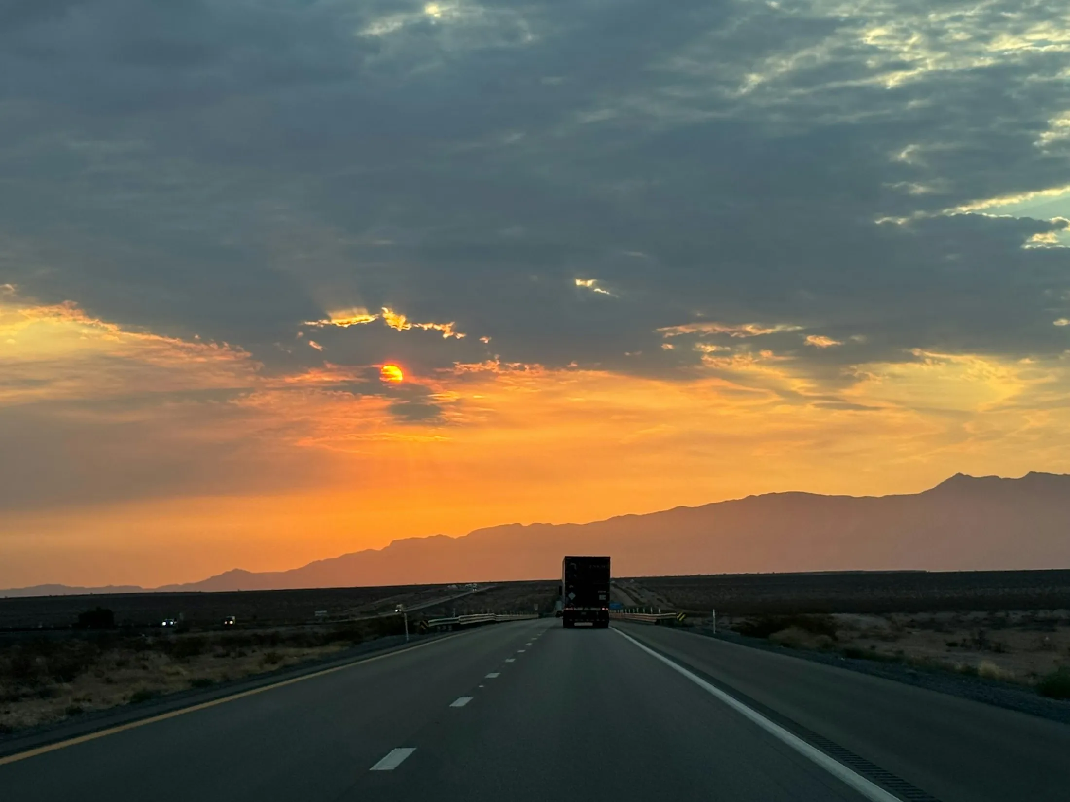 A truck on an open highway heading towards distant mountains at sunset